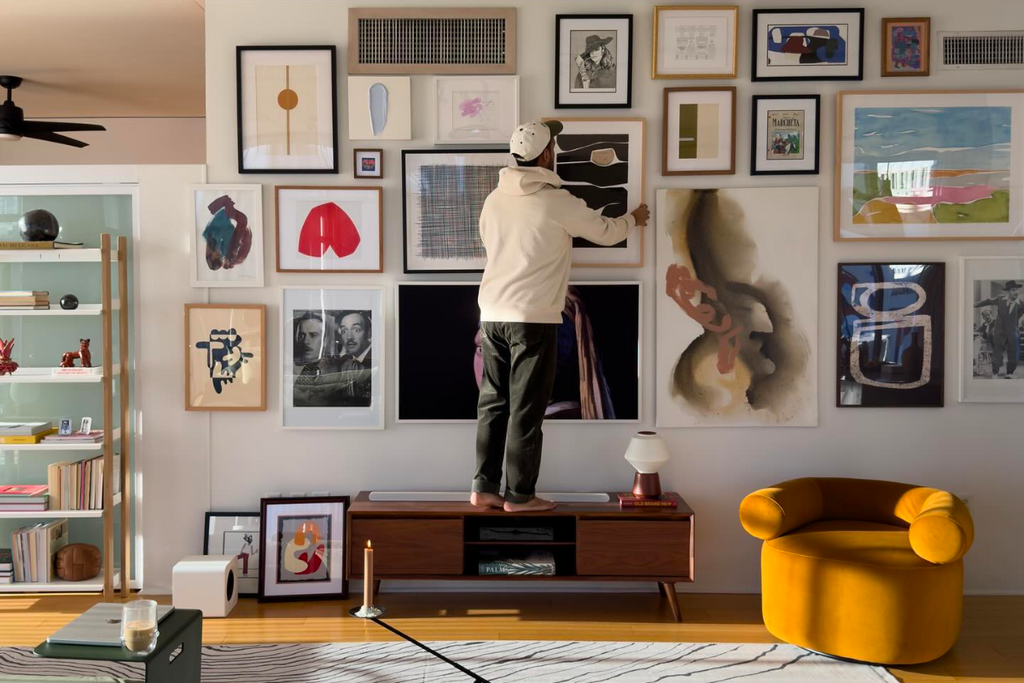 A man wearing a cream hoodie and black jeans is standing on top of a wooden console. He is adjusting an artwork situated in the centre of a gallery wall in his apartment.