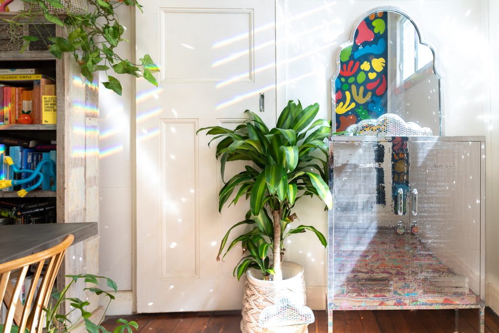 A Midi locker is covered in mirrored squares so that it looks like a square mirrorball. It sits in a white room with leafy plants, and there are glittery shards of sunlight scattering off its reflection.