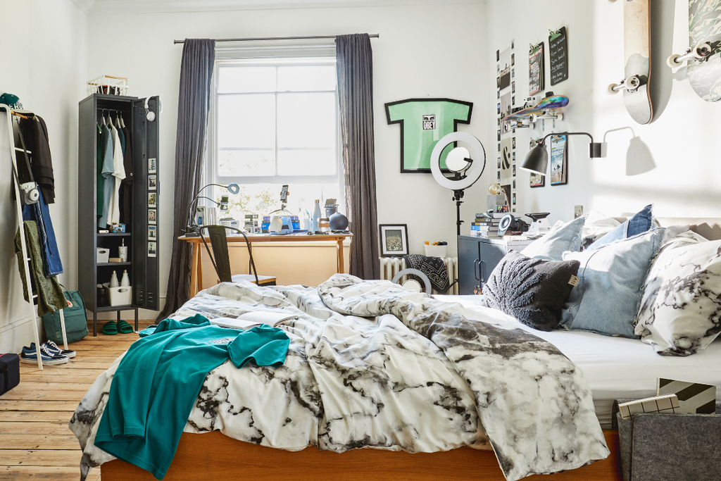 A large, brightly lit room shows a roughly made bed in front of a window and a Skinny locker with the door open. There are posters on the wall and eclectic decorations on a table.